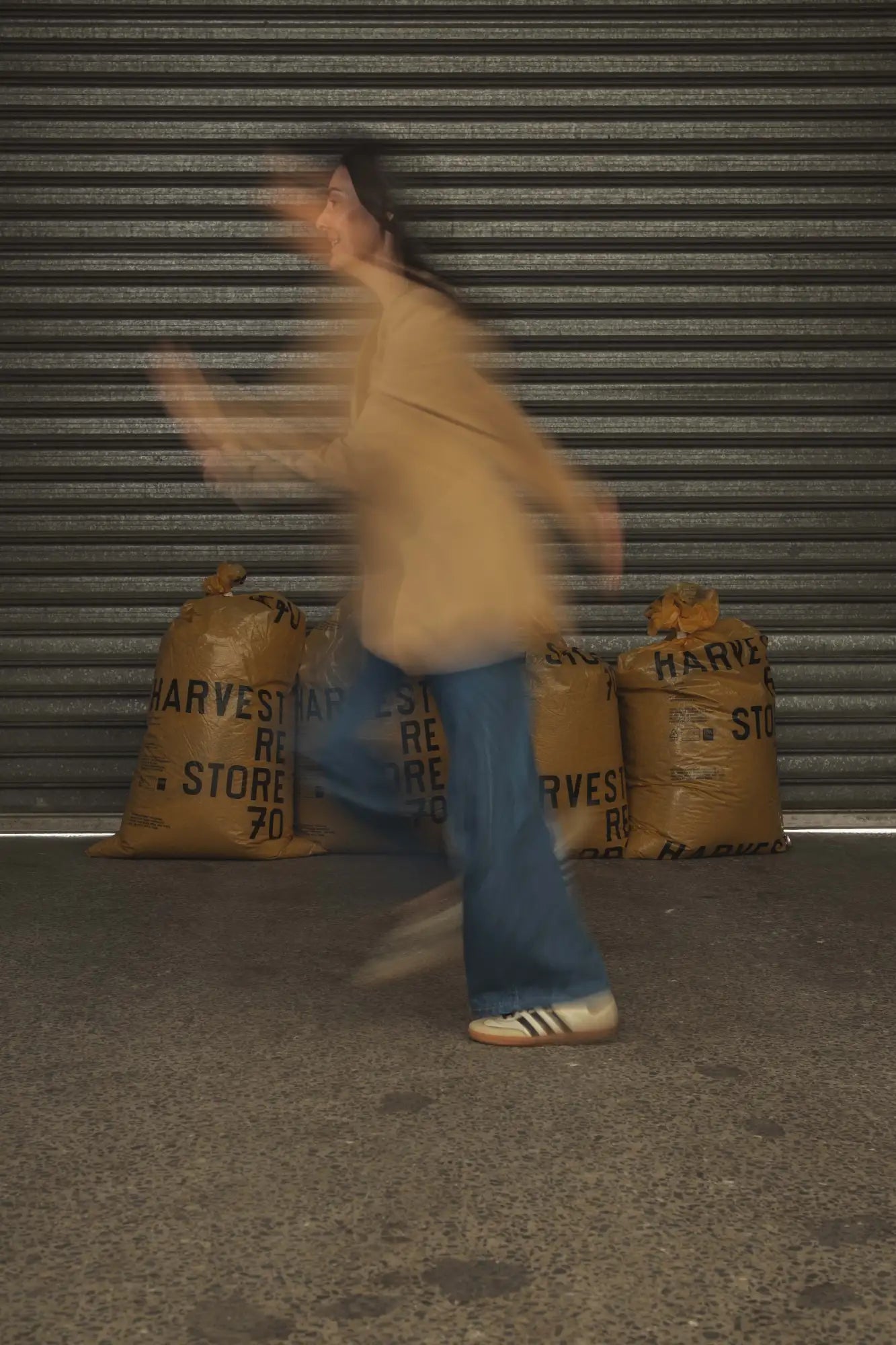A person in motion wearing a tan oversized sweater, blue flared jeans, and beige adidas sneakers with three black stripes.