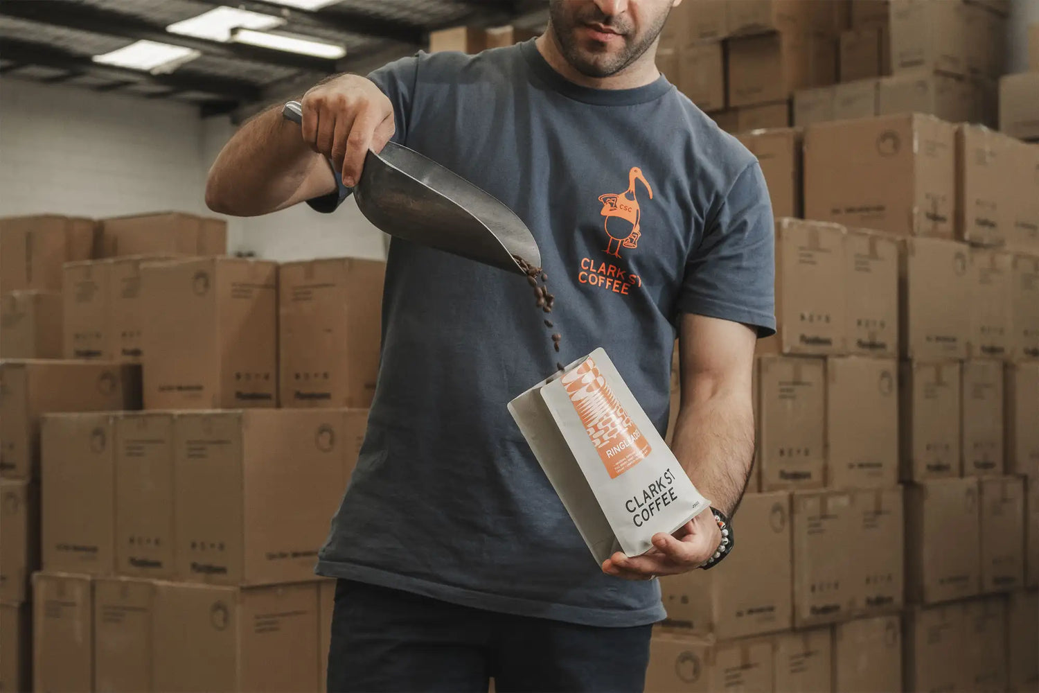 A sleek, rectangular white coffee bag with orange branding and text reading ’clark’s coffee’ is being filled from a metal scoop by a man in a warehouse setting.