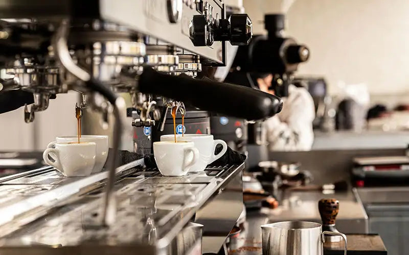 White ceramic espresso cups sit beneath a professional coffee machine as rich, dark liquid pours into them.